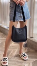 Model carrying the Herme$ Black Taurillon Clemence Leather Evelyne III PM with gold hardware