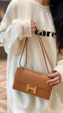 Model carrying the brown Epsom Herme$ Constance Long Wallet To Go with its shoulder strap