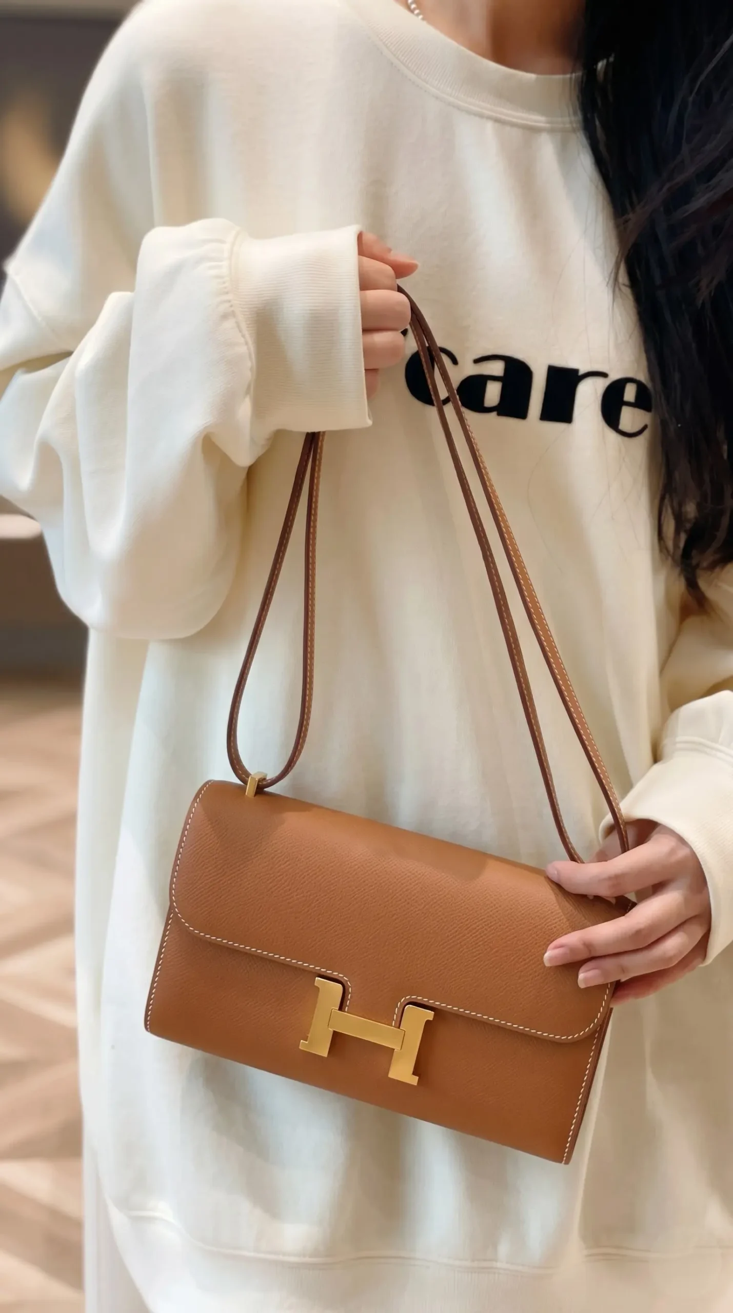 Model carrying the brown Epsom Herme$ Constance Long Wallet To Go with its shoulder strap
